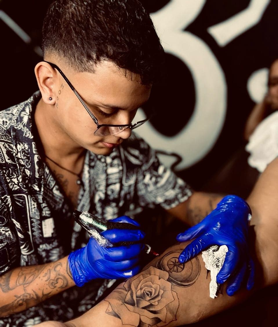 Javier Mena tatuando en el estudio, con guantes y la máquina en la mano, trabajando una rosa en el antebrazo de un cliente