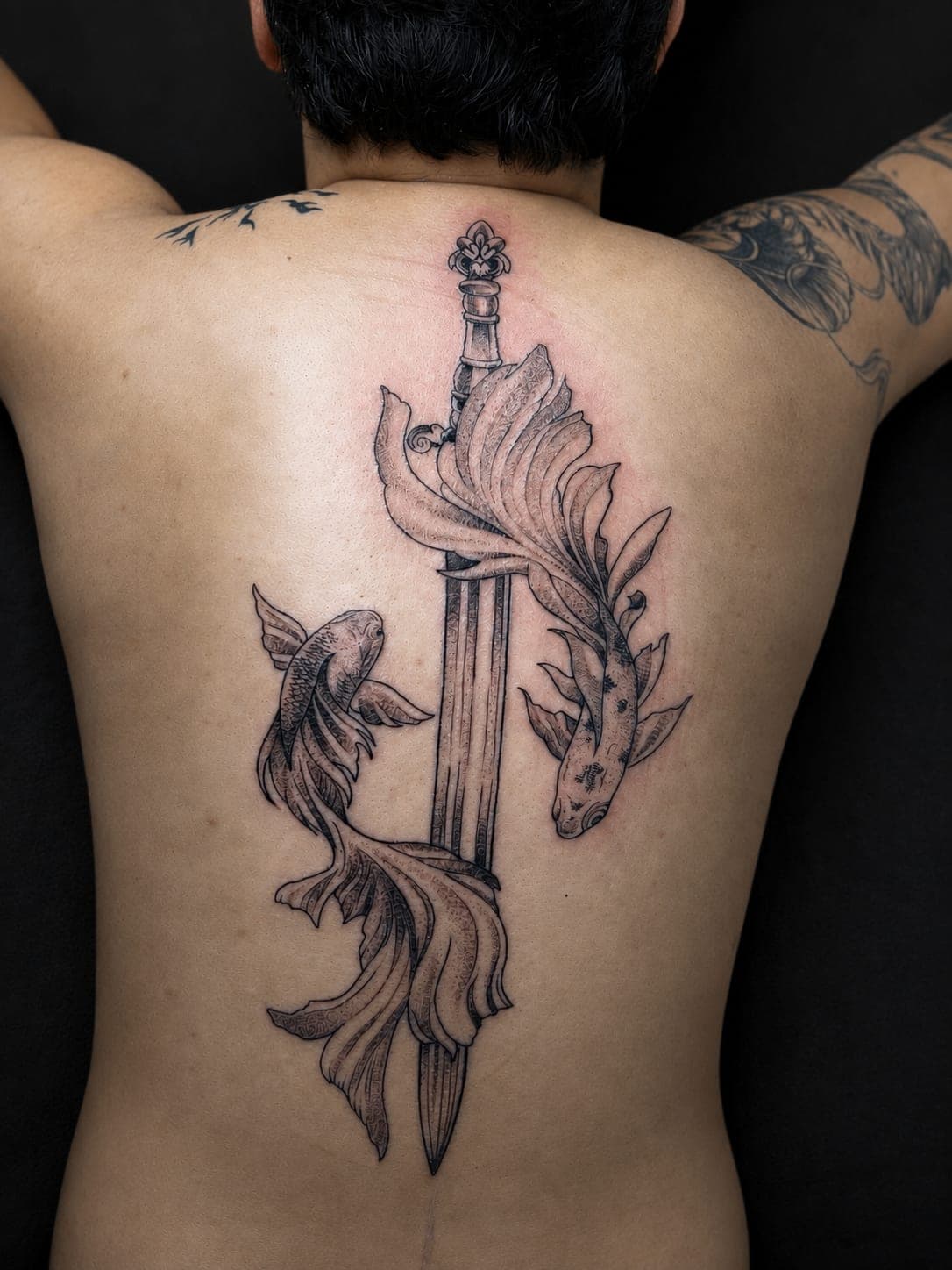 Dos peces betta enroscados en una espada clavada, tatuados a lo largo de la columna en línea negra con sombreado suave